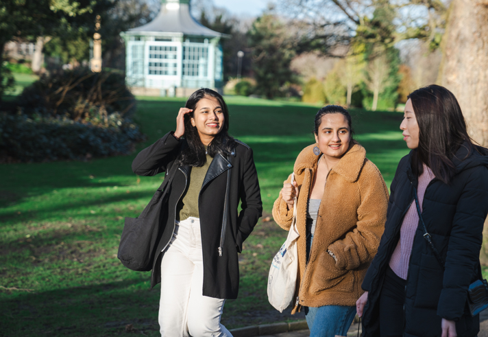 Three people walking together along a paved path in a park with green grass and trees. They are dressed in warm, casual clothing, including coats and jackets, and carrying bags. In the background, there is a light blue gazebo surrounded by greenery, and the scene is lit by natural daylight.