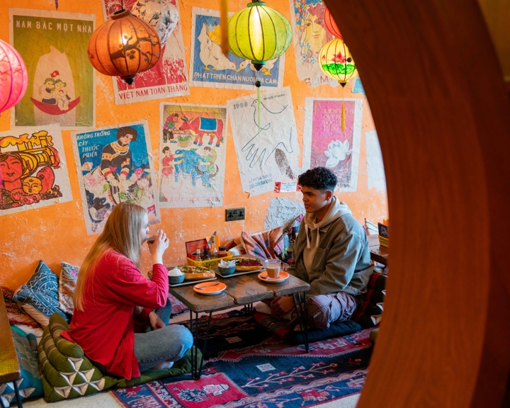 A cozy café interior with vibrant orange walls decorated with colorful Vietnamese posters and artwork. Several hanging lanterns in red, yellow, and green provide warm lighting. Two people are seated on floor cushions at a low wooden table, enjoying food and drinks. The table holds plates and cups, and the floor is covered with patterned rugs, creating a relaxed, intimate atmosphere.