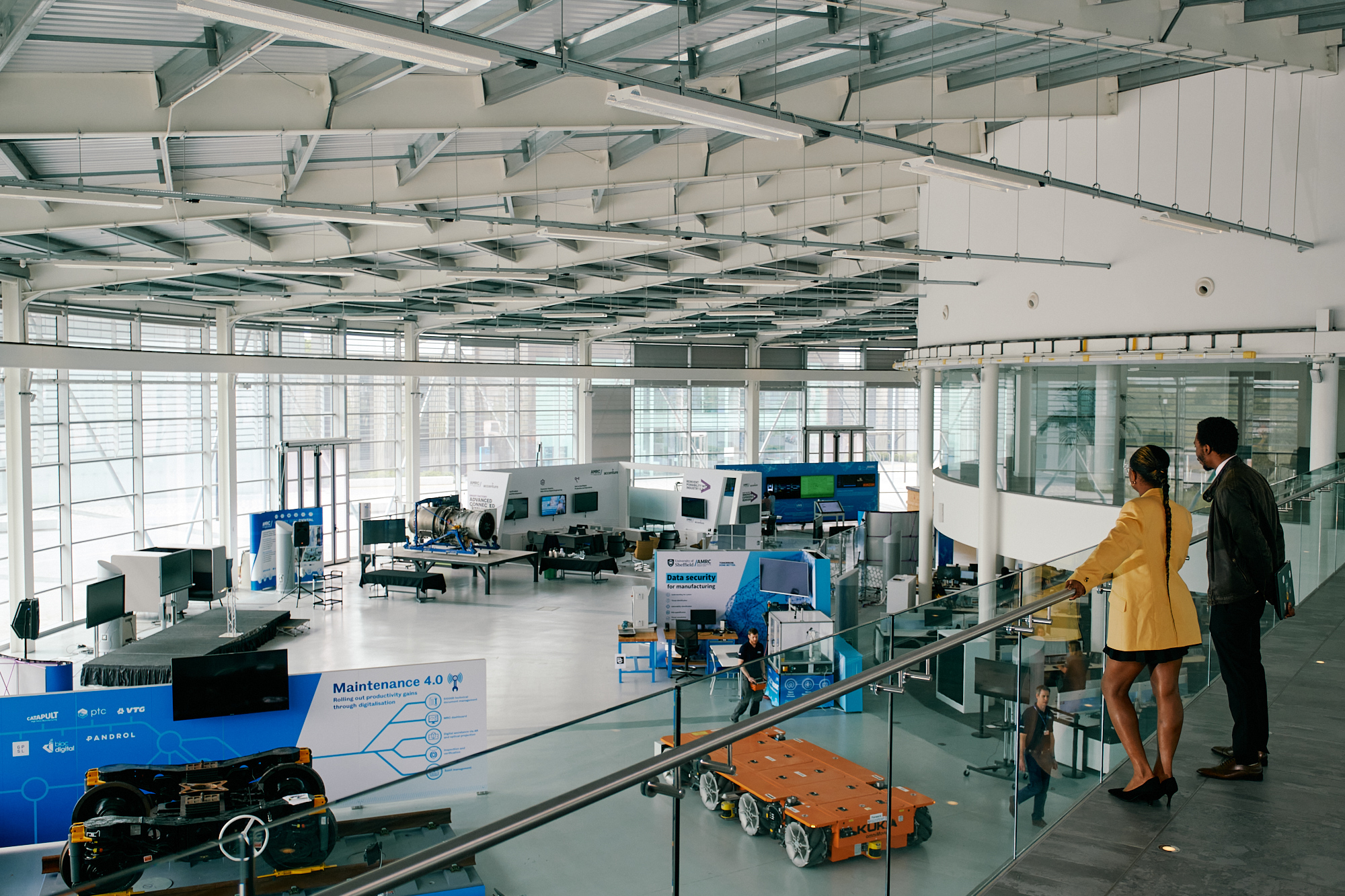 Interior view of a modern industrial facility with high ceilings and large windows. The space contains advanced machinery, robotic platforms, and technology displays, including a blue sign labelled ‘Maintenance 4.0.’ Two people stand on an upper-level glass balcony overlooking the equipment below.