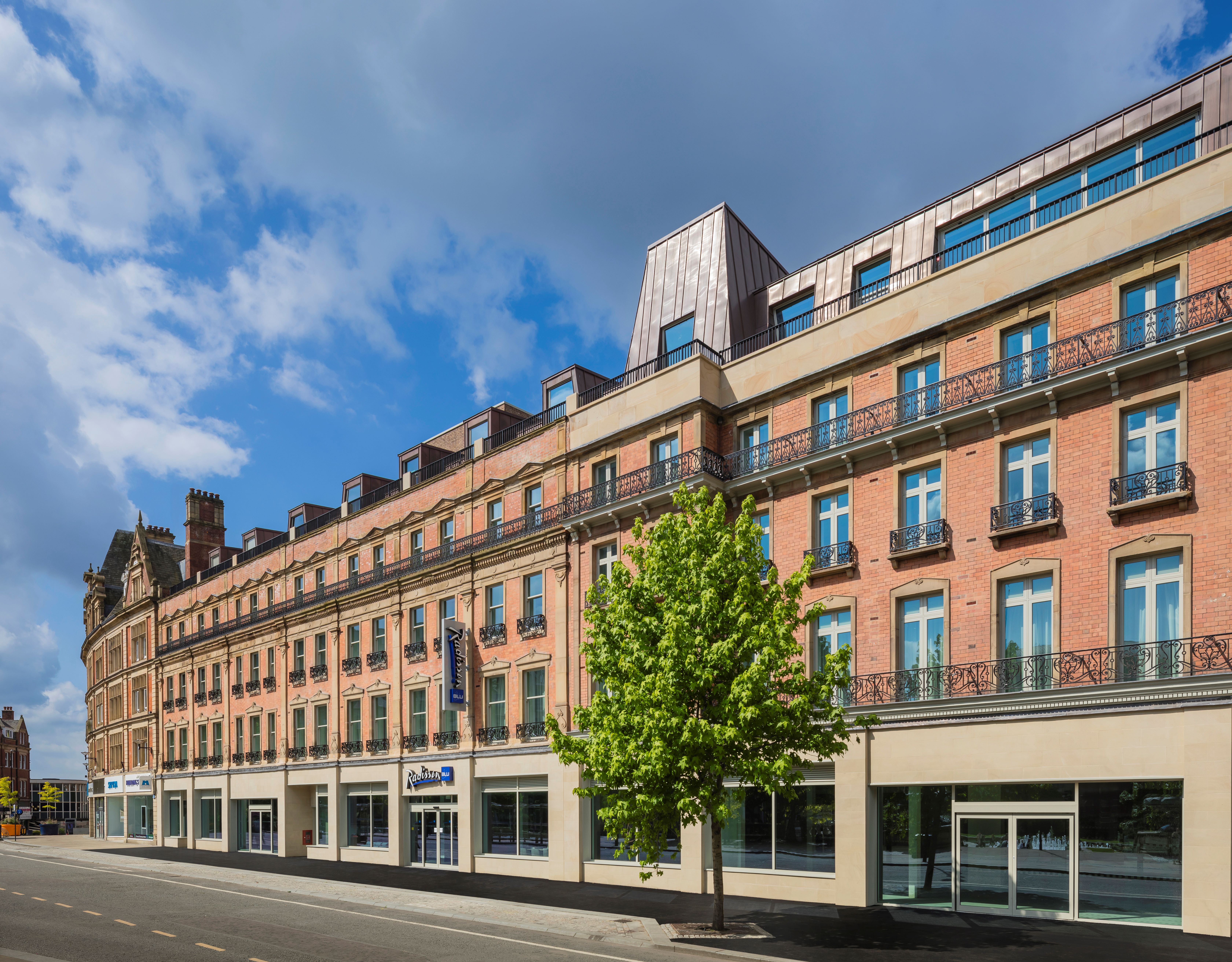 CGI-rendering of a Street view of a large, multi-story building with a red-brick façade and beige stone accents, featuring rows of tall windows with black wrought-iron balconies. The ground floor has several glass storefronts, including a visible sign for ‘Travelodge.’ A single green tree stands on the sidewalk in front of the building, and the sky above is partly cloudy with patches of blue.