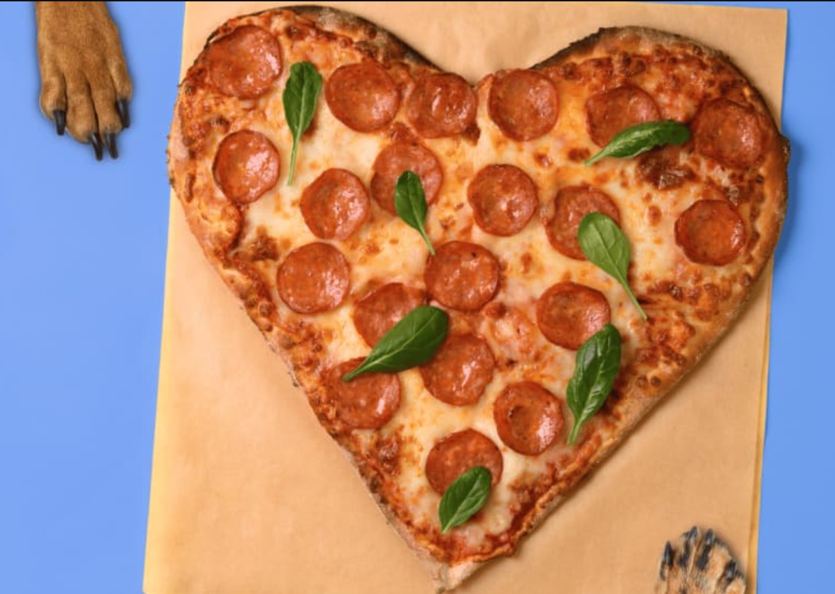 A pizza, in a heart-shape, sits on a wooden cutting board. Just next to the pizza is a dog's paw.