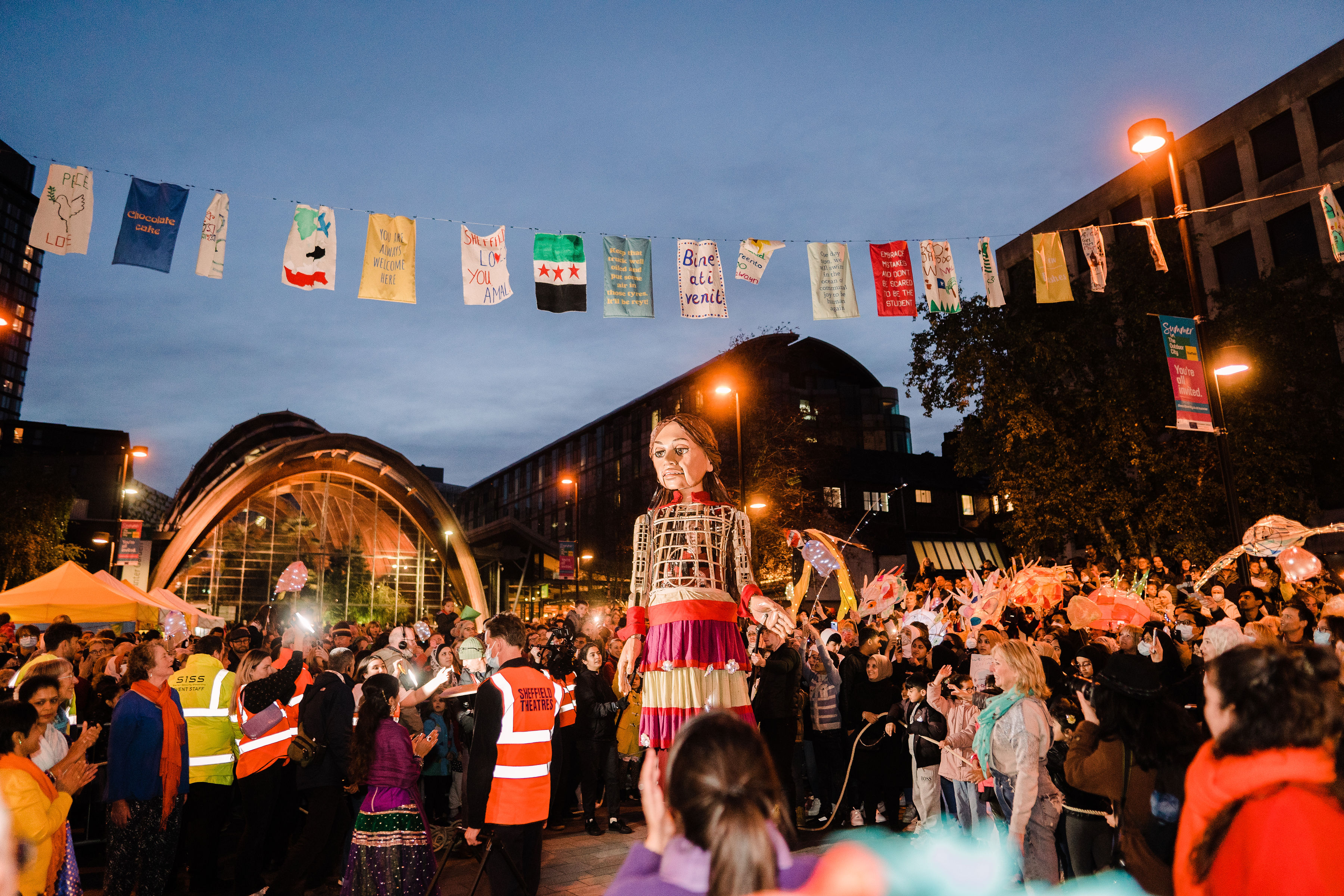 Outdoor evening street festival with a large crowd gathered around performers. Colorful flags hang overhead, and event staff in orange vests are visible. A tall figure in a decorative costume stands in the centre, with illuminated buildings and a glass arch structure in the background.