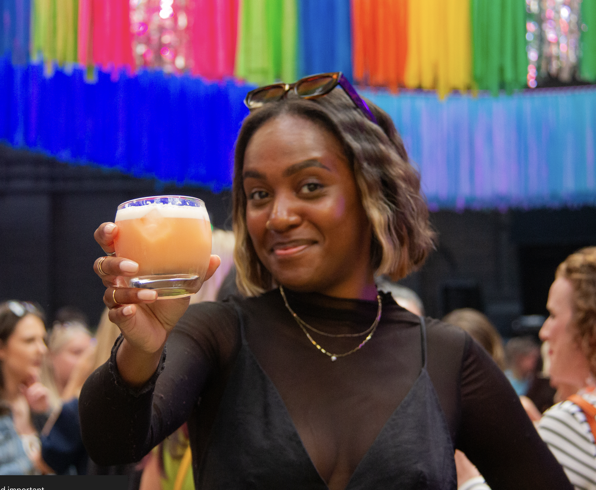 A person at a lively event holds up a glass of orange‑coloured drink as if making a toast. They wear dark, stylish clothing with layered necklaces and sunglasses resting on their head. Behind them, colourful hanging decorations in bright rainbow shades fill the space, with a crowd of people gathered in the background.