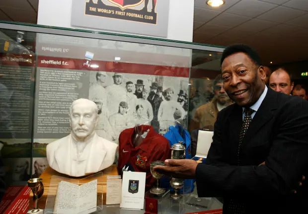 Display case featuring Sheffield FC memorabilia, including a white bust, vintage football shirts in red and blue, silver trophies, and historical documents. A black-and-white photograph of early football players is mounted in the background, with signage reading ‘The World’s First Football Club.’
