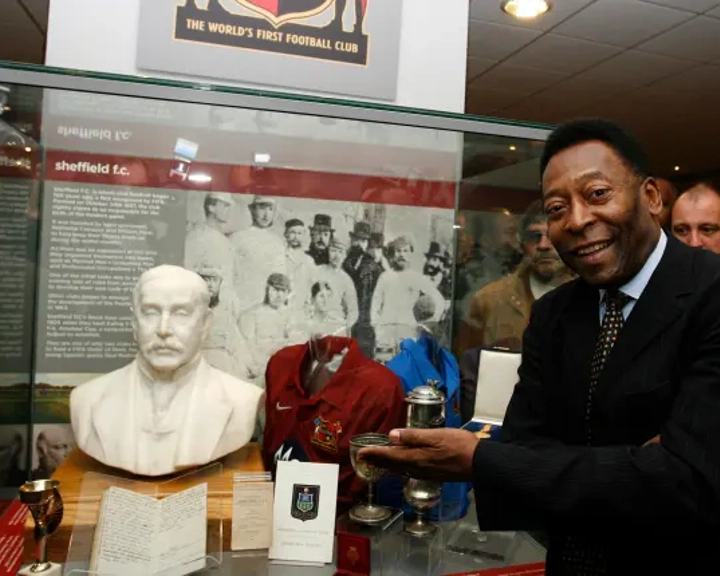 Display case featuring Sheffield FC memorabilia, including a white bust, vintage football shirts in red and blue, silver trophies, and historical documents. A black-and-white photograph of early football players is mounted in the background, with signage reading ‘The World’s First Football Club.’