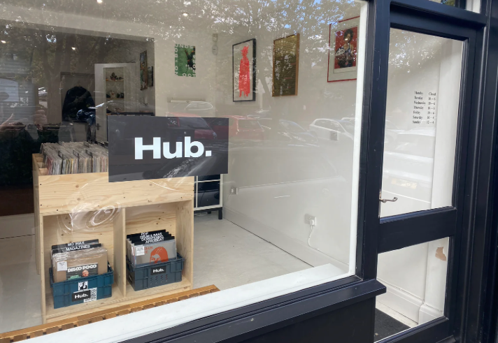 The window of a record store with a sign in the window that reads 'HUB.' . In the shop you can see wooden racks filled with vinyl records.