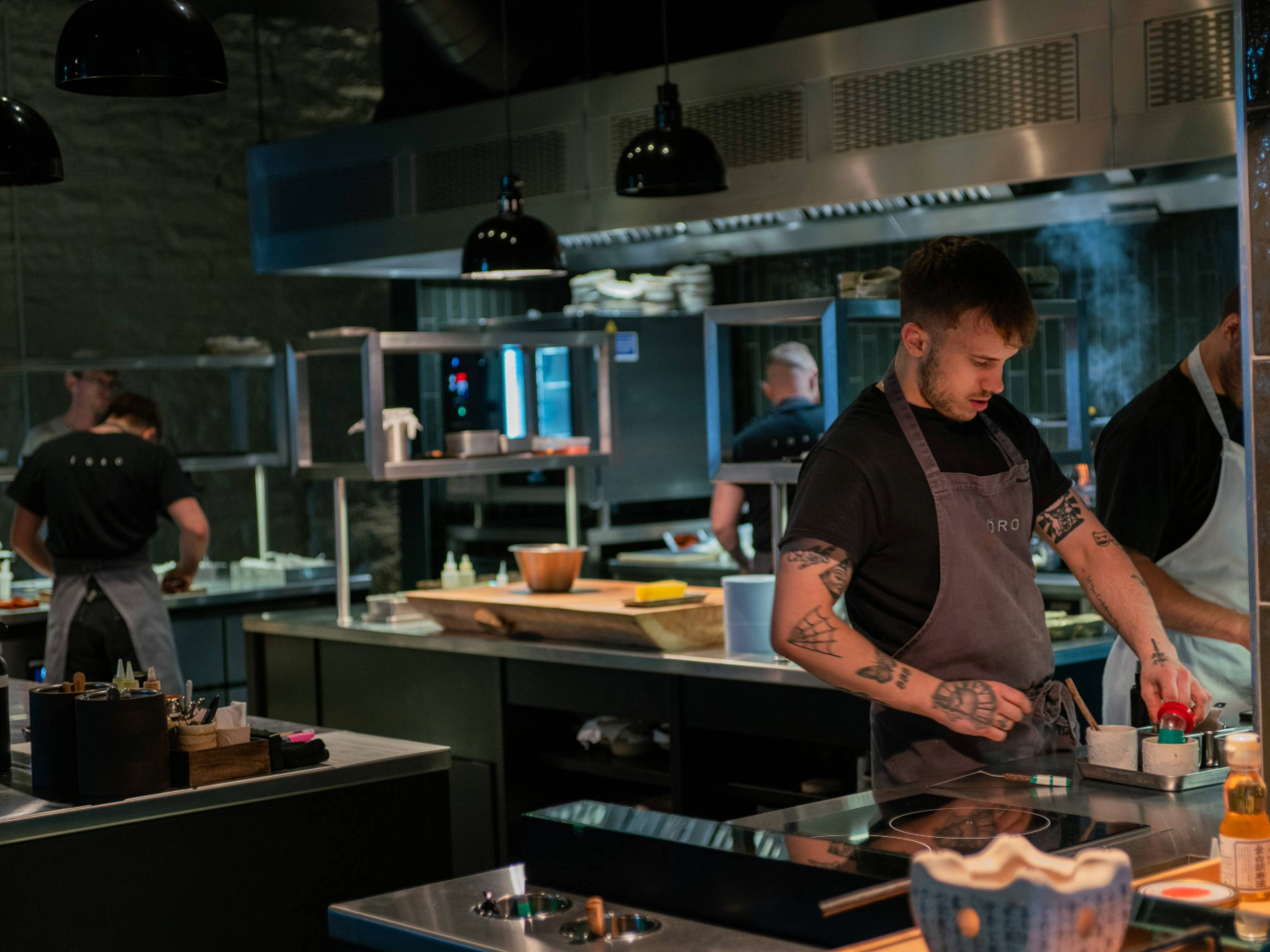 Chefs working in a modern open kitchen with stainless steel counters and cooking stations. One chef in the foreground, wearing a grey apron, is preparing food near an induction hob with small bowls and utensils. The background shows other chefs at work, shelves with plates, and a large oven. Black pendant lights hang from the ceiling, and the setting has an industrial, contemporary design.