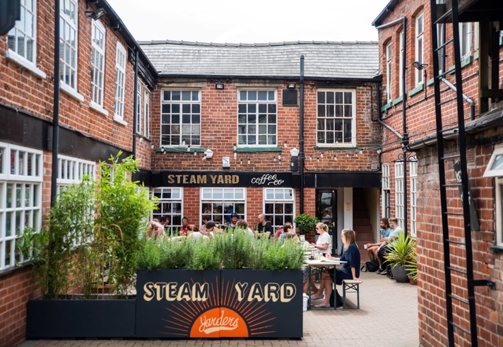 Outdoor courtyard of Steam Yard café with people sitting at tables, surrounded by red‑brick buildings and greenery.