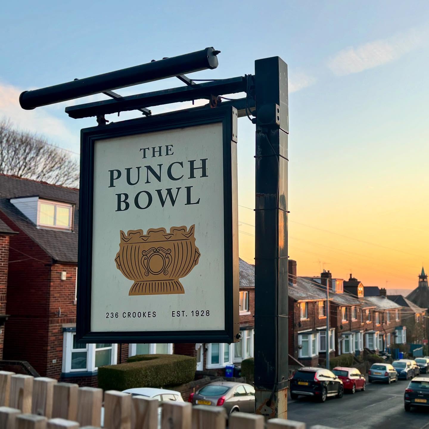 A hanging pub sign for The Punch Bowl in Crookes, featuring a stylised punch bowl illustration, photographed at sunset with rows of terraced houses in the background.