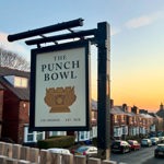 A hanging pub sign for The Punch Bowl in Crookes, featuring a stylised punch bowl illustration, photographed at sunset with rows of terraced houses in the background.