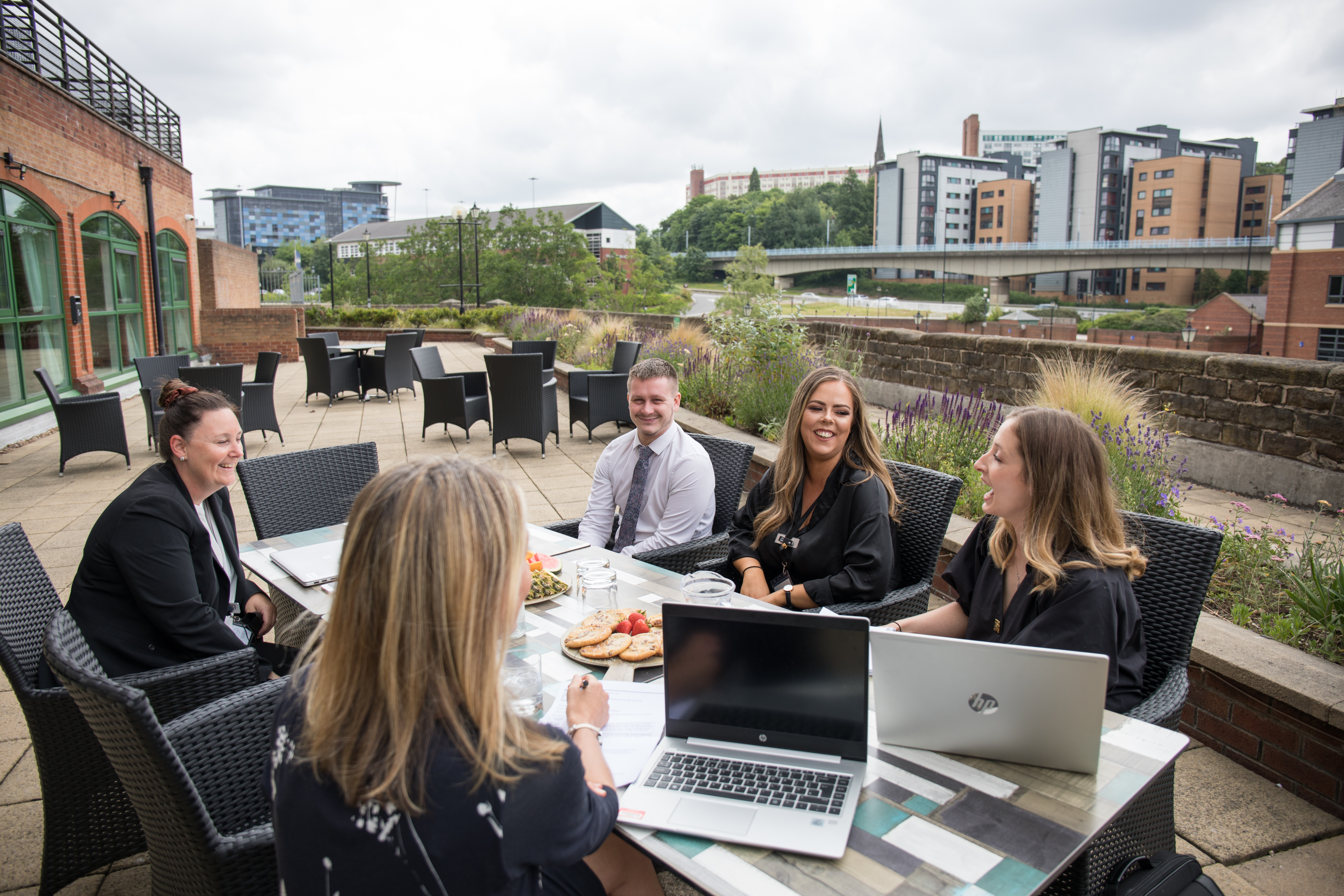 Five people, of varied ages and appearances, sit around a table on an outdoor patio, engaged in conversation and collaborative work. The table includes open laptops, notebooks, and a plate of snacks. The setting is wheelchair accessible and features a cityscape backdrop with modern buildings and greenery, providing a relaxed and inclusive environment.