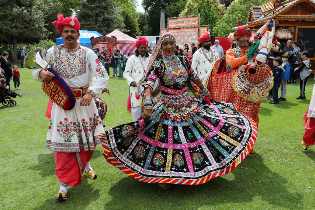  A vibrant outdoor performance featuring dancers in traditional Indian attire. The central dancer wears an ornate, multi-coloured dress with intricate patterns and flared skirt, surrounded by performers in embroidered white and red costumes. One performer holds a decorated horse prop. Behind them, festival stalls and onlookers create a lively backdrop on a grassy area with trees.
