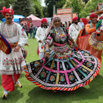  A vibrant outdoor performance featuring dancers in traditional Indian attire. The central dancer wears an ornate, multi-coloured dress with intricate patterns and flared skirt, surrounded by performers in embroidered white and red costumes. One performer holds a decorated horse prop. Behind them, festival stalls and onlookers create a lively backdrop on a grassy area with trees.