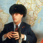 Three children sit behind microphones at a model international conference table in front of a large world map. Each child wears distinctive headwear representing different cultures or countries, with small national flags placed on the table. The central child wears a dark suit and large fur hat, while the others wear a sombrero and a wrapped turban-style head covering. The scene appears staged as part of a school presentation or mock diplomatic event.