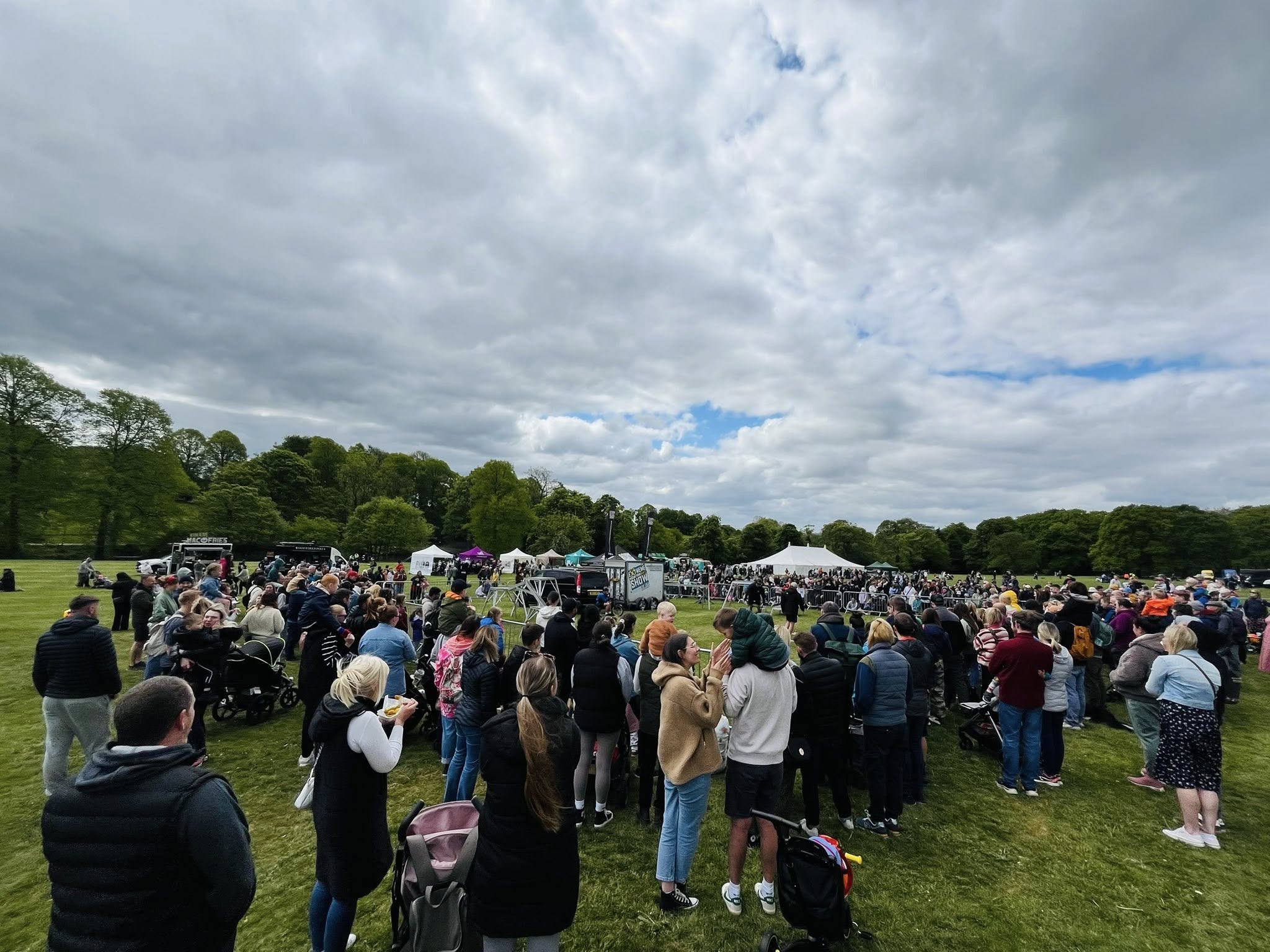 A large crowd gathered on a grassy field at an outdoor event, with people standing and watching activities near white and green tents in the distance. Many attendees are wearing coats and jackets, and some have pushchairs. The background shows trees lining the field under a mostly cloudy sky with patches of blue. The scene suggests a lively community gathering or fair.