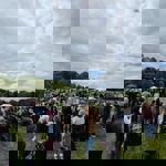 A large crowd gathered on a grassy field at an outdoor event, with people standing and watching activities near white and green tents in the distance. Many attendees are wearing coats and jackets, and some have pushchairs. The background shows trees lining the field under a mostly cloudy sky with patches of blue. The scene suggests a lively community gathering or fair.
