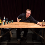 A man sat at a large wooden table, with various blocks, bottles, cans, and small boxes arranged on the surface of the table.