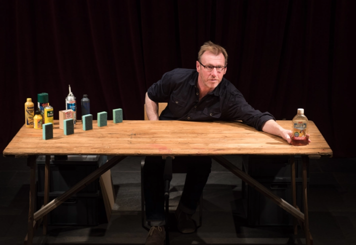 A man sat at a large wooden table, with various blocks, bottles, cans, and small boxes arranged on the surface of the table.