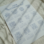 A single-page 2026 calendar printed in blue ink on white paper, featuring hand-drawn floral illustrations for each month. The calendar is laid flat on a light fabric surface with soft shadows cast across it.