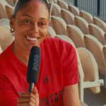Person seated among rows of empty stadium seats holds a handheld microphone toward the camera. Wearing a bright red sports shirt with a small black logo, the person is positioned in the foreground while curved beige seats fill the background. Sunlight illuminates the stand, creating a warm atmosphere and highlighting the repeating pattern of seats stretching across the image.