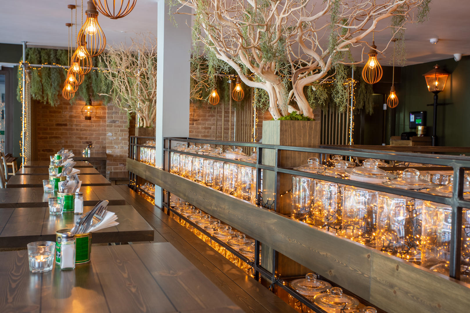 A stylish restaurant interior with long wooden tables, warm pendant lights, decorative trees in large planters and shelves of glass jars lit with fairy lights, creating a soft, ambient glow.