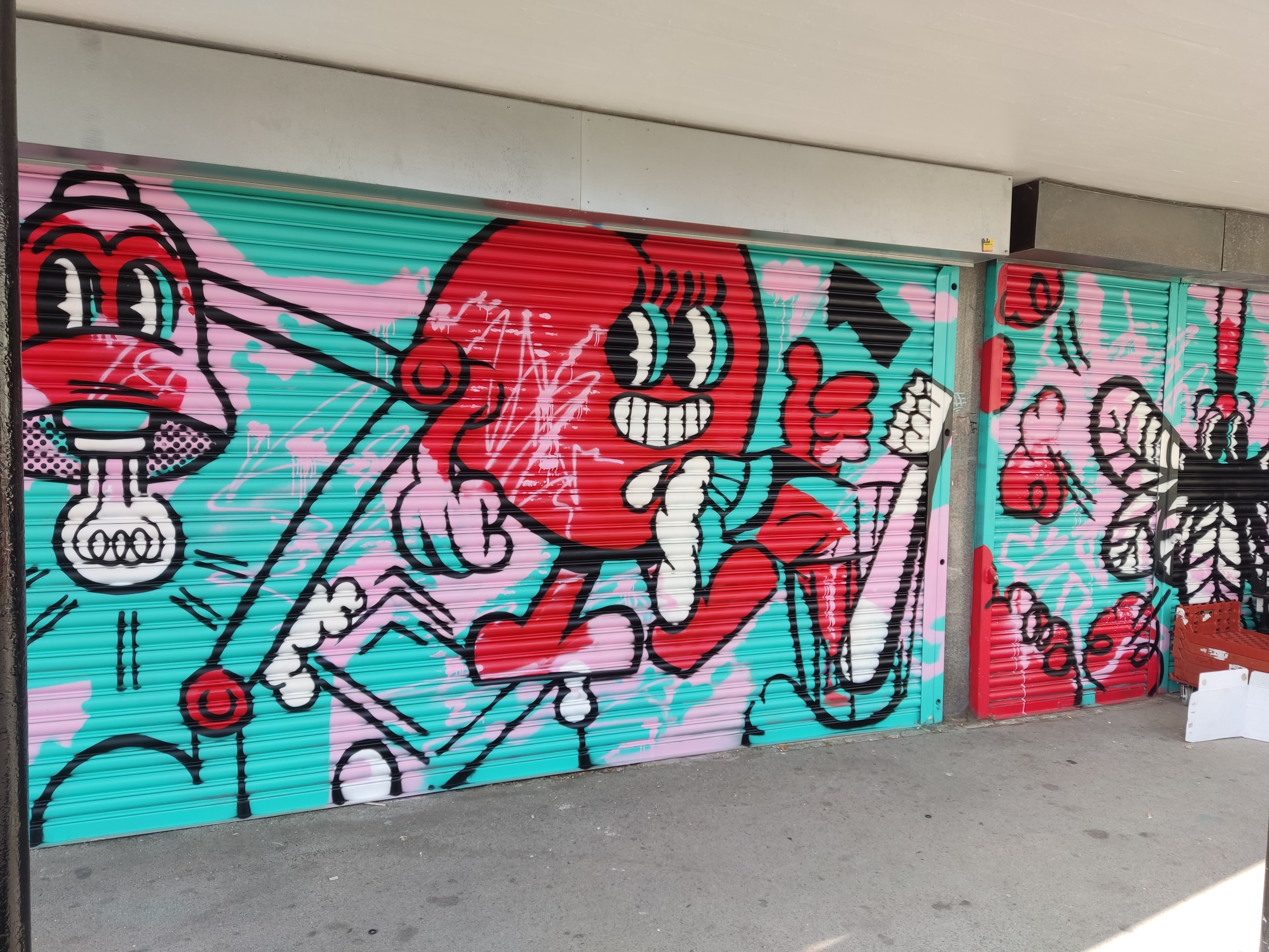 Colorful street art painted on metal shutters featuring bold cartoon-style characters in red, black, and white against a turquoise and pink background. The central figure is a smiling red character with exaggerated eyes and limbs, surrounded by abstract shapes and other stylized faces.