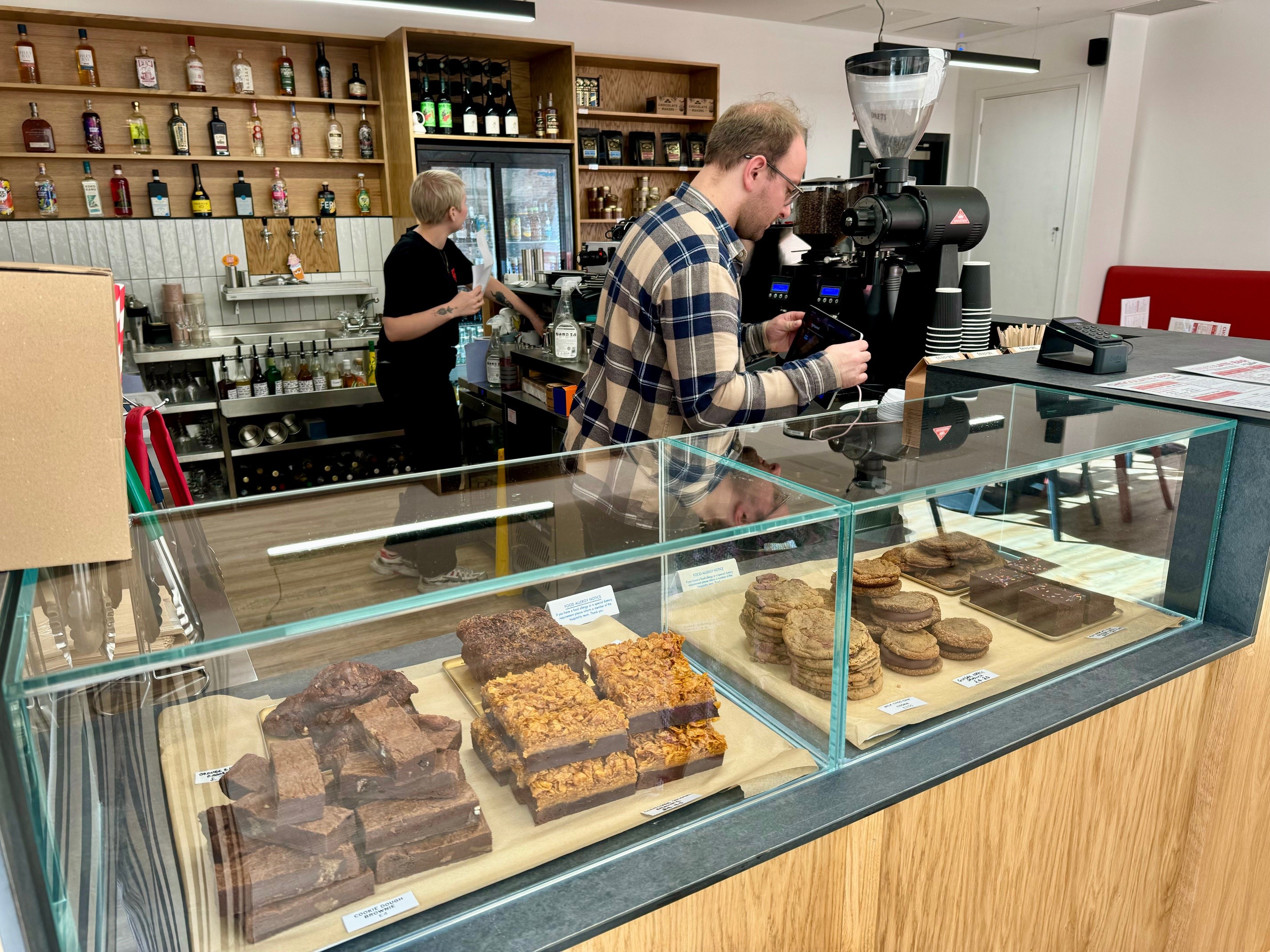 Interior of a café with a wooden counter and glass display case filled with assorted baked goods, including brownies, flapjacks, and cookies. Behind the counter, shelves stocked with bottles and coffee-making equipment are visible, along with a large espresso machine.