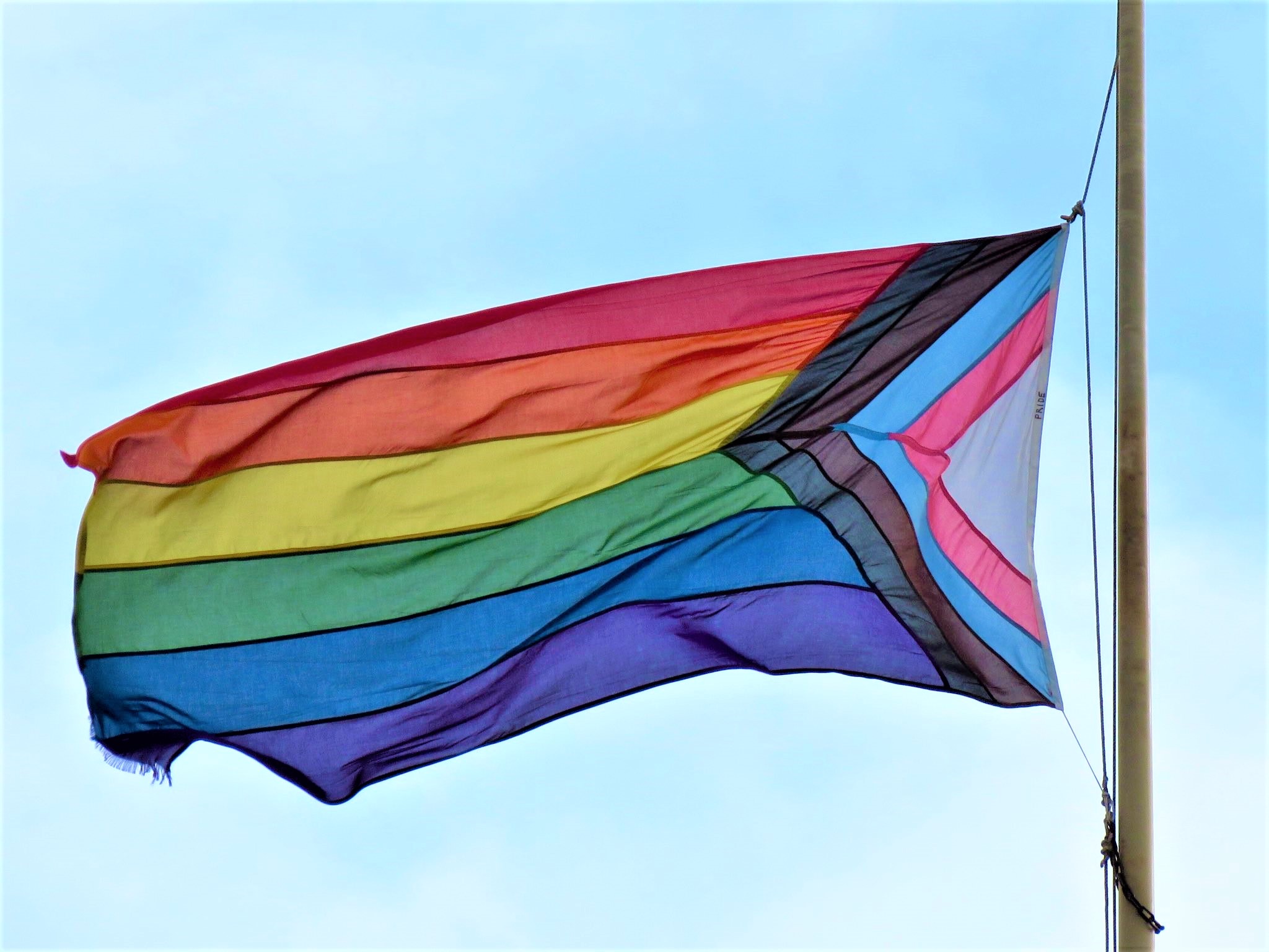 Progress Pride flag flying on a flagpole against a clear blue sky. The flag features horizontal rainbow stripes along with a chevron design in black, brown, white, pink, light blue, and blue.