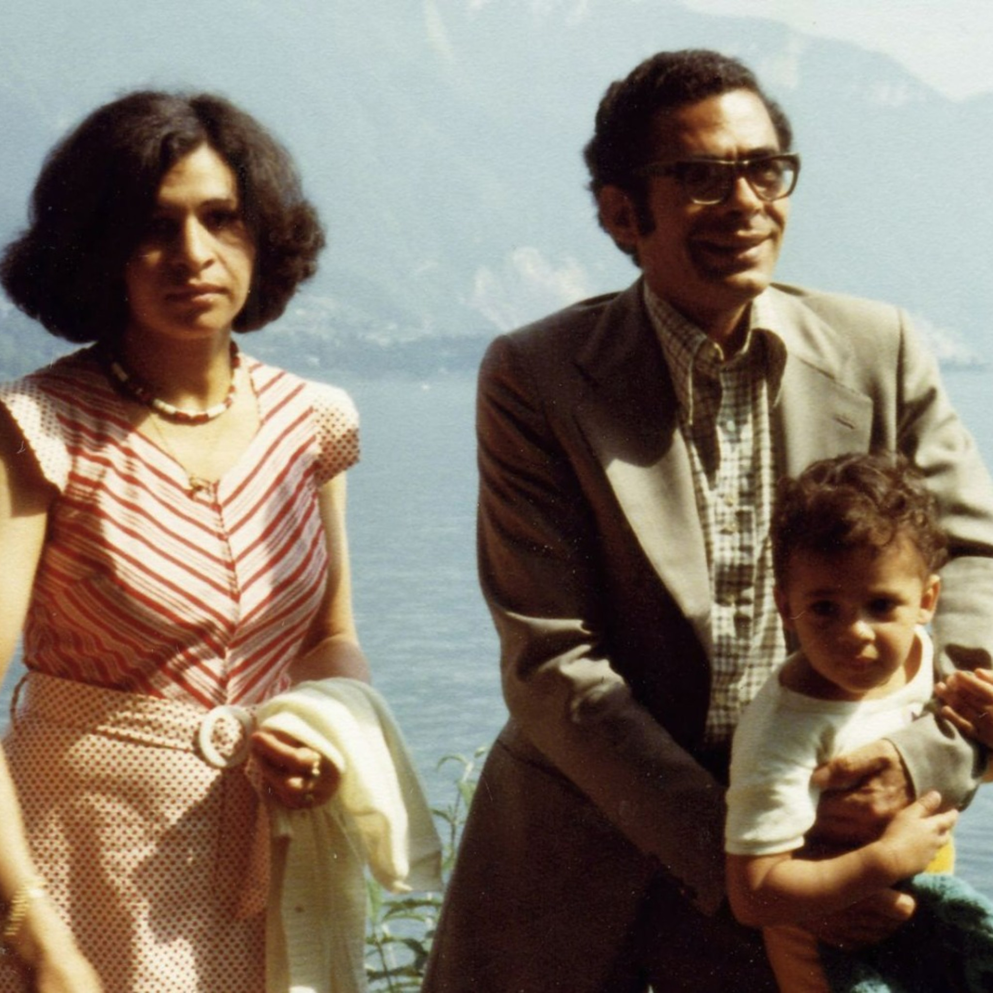Vintage family photograph showing three people outdoors beside a large body of water. One adult in a patterned sleeveless dress stands on the left holding a light-coloured garment. Another adult in a jacket, checked shirt and large glasses stands on the right, holding a young child. The background shows a lake and distant mountains under a bright sky. The image has the soft colours and slight fading typical of an older printed photograph.