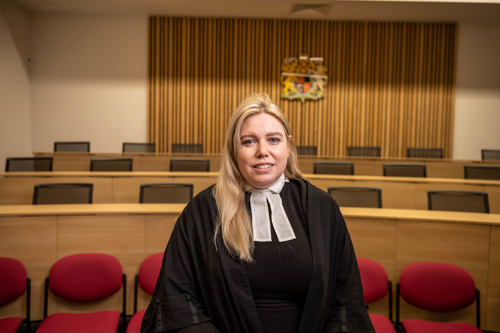 A person wearing a black robe and white neck bands is standing in a courtroom setting. Behind them are rows of red chairs and wooden benches, with a wooden panel wall featuring a coat of arms at the center. The lighting is warm, and the room has a formal, official atmosphere.