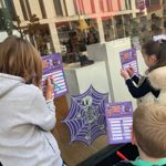 Three children stand outside a shoe shop window, completing purple Halloween Trick or Treat Trail activity sheets with coloured pens. A large skeleton on a spiderweb decoration is displayed in the window, alongside boots and shoes. Reflections of nearby buildings appear in the glass. A prominent red sale sign shows “£38”, and other sale notices are visible inside the shop. The children appear to be taking part in a Halloween-themed town-centre trail.