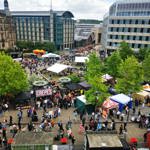 A wide aerial view of a food festival with colourful stalls, trees, and surrounding city buildings.
