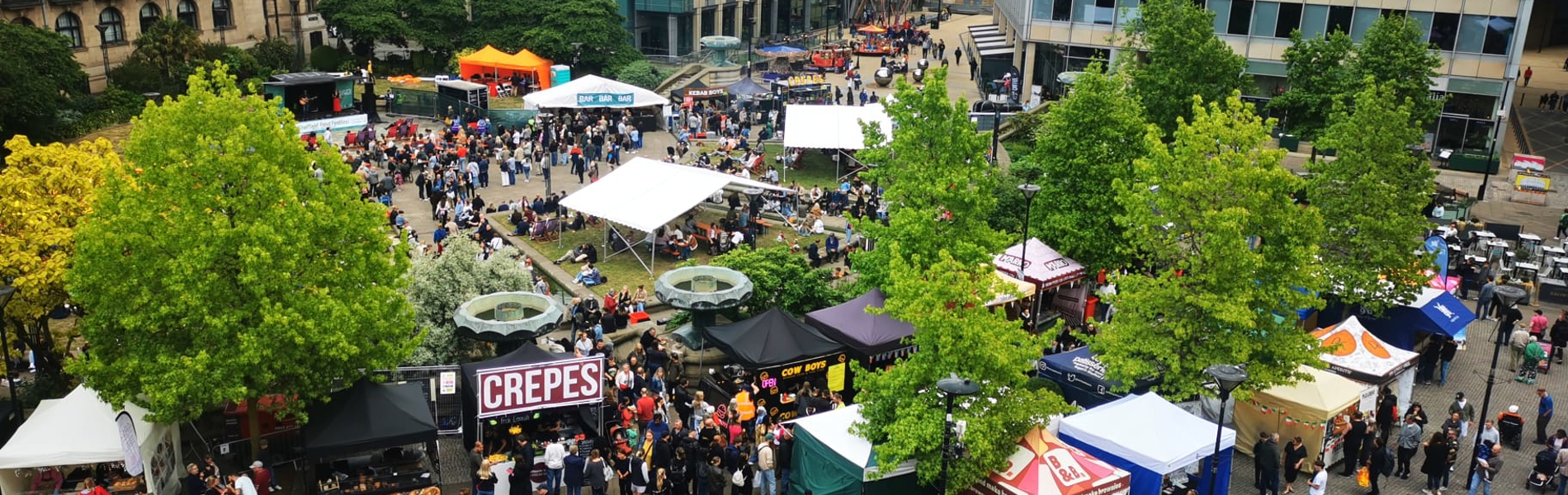 A wide aerial view of a food festival with colourful stalls, trees, and surrounding city buildings.