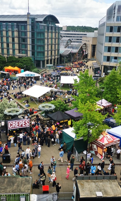 A wide aerial view of a food festival with colourful stalls, trees, and surrounding city buildings.