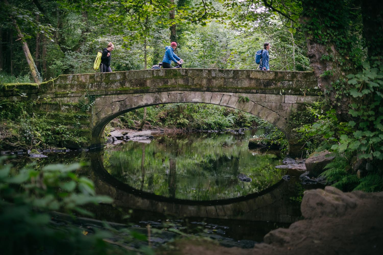 Three people are crossing a stone bridge over a stream in a forest.