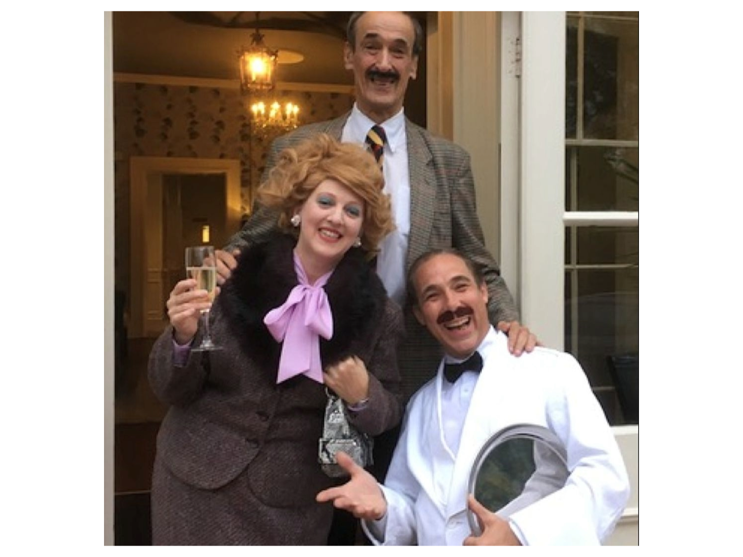 Three people are standing in front of an open doorway with glass panels. The person on the left is wearing a brown textured suit with a large black fur collar and a bright pink bow tie, holding a champagne flute in one hand and a handbag in the other. The person in the middle is dressed in a grey checked blazer with a white shirt and striped tie. The person on the right is wearing a white tuxedo jacket with a black bow tie and is holding a silver serving tray. 