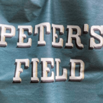 Poster artwork featuring a close-up section of a football-style banner in teal-blue fabric with large white three-dimensional lettering reading “PETER’S FIELD”. The banner appears slightly folded, creating shadows across the text. At the bottom, the name “Sean Cooney” is printed in small white text on a black strip. The image has a textured, printed-poster appearance.