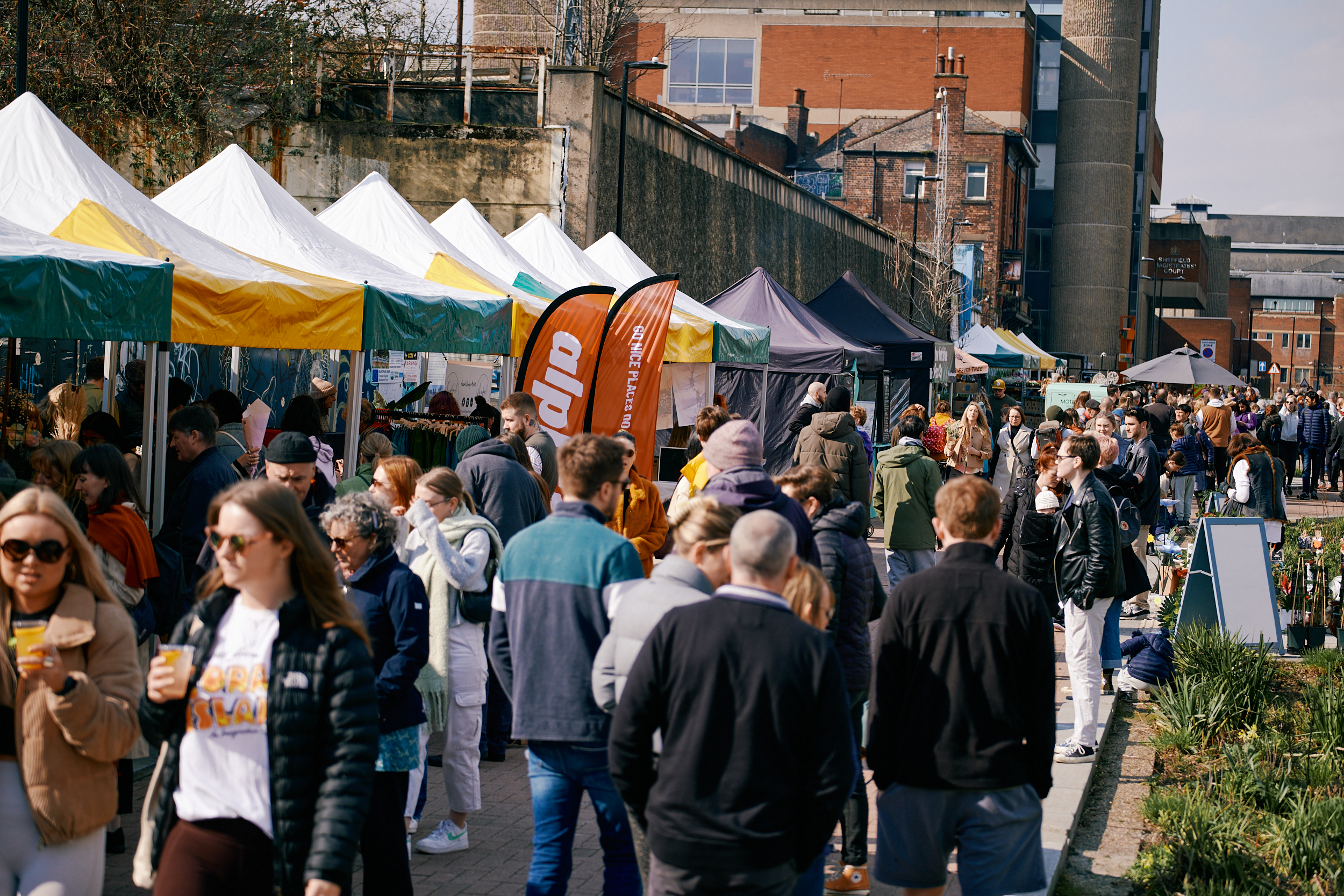 An open air market in a city centre. There are lots of people walking up and down and browsing the stalls.