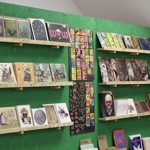 Shelves mounted on a green wall displaying a variety of illustrated prints and artwork, featuring bold colours, detailed drawings, and creative designs in different sizes.