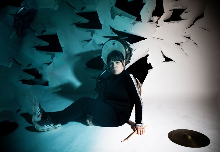 A person is sat on the floor in an empty white room. They have a drum stick in their hand, there is a snare drum behind their head and a cymbal lies on the floor to their right. The wall behind them is damaged, with multiple holes/ tears in the fabric of the surface.