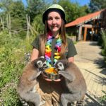 A zoo keeper stands on a sunny garden path holding a bowl of fruit while two ring-tailed lemurs cling to her arms and eat from the bowl. She wears a colourful bucket hat and rainbow flower garland. Lush greenery and a building are visible in the background, creating a bright outdoor wildlife park setting.