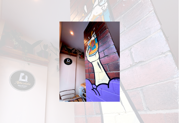 Interior view of Abbeydale Beerworks Taproom. A painted mural covers a red brick pillar, showing a large hand holding a foaming beer glass filled with colourful illustrations of books, buildings and other objects. The lower section of the pillar is painted purple. In the background are wooden shelves with potted plants, metal-framed chairs, and an Abbeydale Brewery wall sign. Warm ceiling lighting illuminates the modern taproom space.