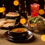 A close-up of an assortment of dishes on a wooden table, including a bowl of curry, rice, flatbreads, a spiced vegetable dish and two colourful cocktails, surrounded by warm blurred lights in the background.
