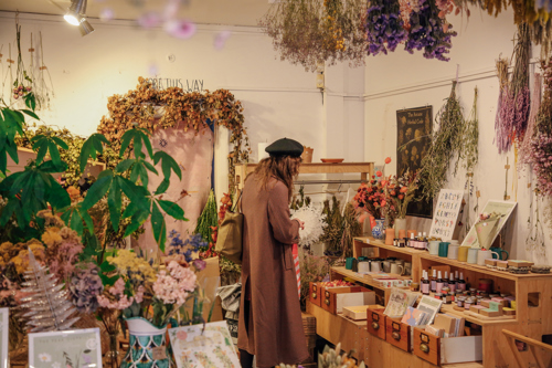 Interior of a cozy shop filled with dried flowers and plants hanging from the ceiling and displayed on shelves. A person wearing a long coat and beret is browsing items on a wooden counter, which holds small jars, books, and decorative pieces. The walls are adorned with floral arrangements and signs, creating a warm, rustic atmosphere.