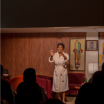 A performer stands barefoot in a warmly lit room set up like a living space, wearing a light-coloured dressing gown and addressing a seated audience. Red armchairs, a side table with a bottle, a floor lamp and framed artwork decorate the wood-panelled backdrop. Audience members appear in silhouette in the foreground, watching the intimate live performance.