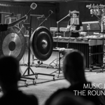 A black and white image of two people on stage performing, surrounded by percussion instruments.