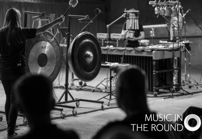 A black and white image of two people on stage performing, surrounded by percussion instruments.