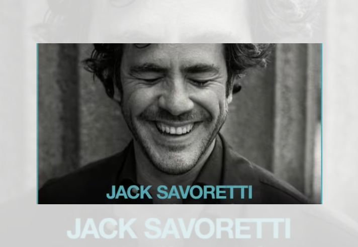 Black-and-white promotional image featuring a close-up portrait of a person with curly hair, eyes closed, and a broad smile. The person wears a dark jacket and is framed against a softly blurred background. At the bottom of the image, the name “JACK SAVORETTI” appears in large light blue capital letters.