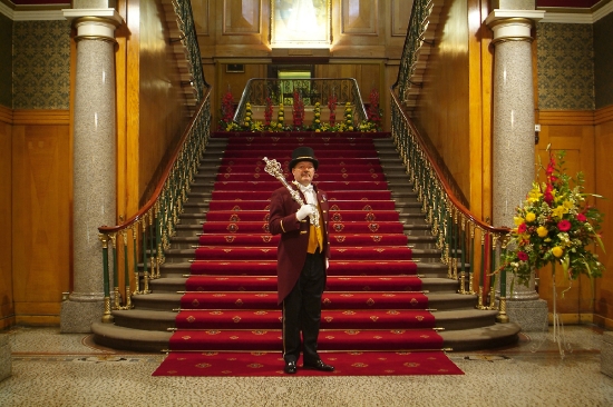 A man stands at the bottom of the main staircase in Cutlers' Hall. He is wearing formal attire and holding a large mace on his shoulder.