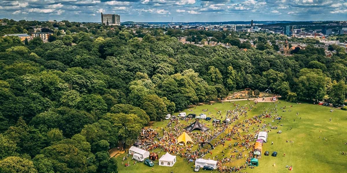 Aerial view of a large outdoor running event in a park, with crowds gathered around tents and a central event area, surrounded by dense trees and the Sheffield skyline in the distance.